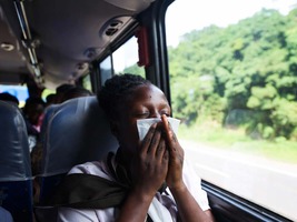 A female passenger properly using an emesis bag to vomit, seated by the window as the bus snakes along the highway through a vast forest. 